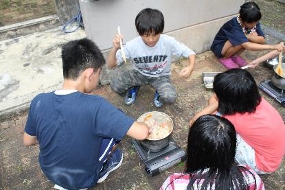 コンロを使って鍋でカレーを煮詰めているのを児童たちが見守っている写真