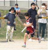 父母が写真を撮る中、児童がゼッケンを纏いバトンを持って走っている写真