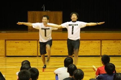 体育館で先生2人がTと書いたシャツを着て、踊っている写真