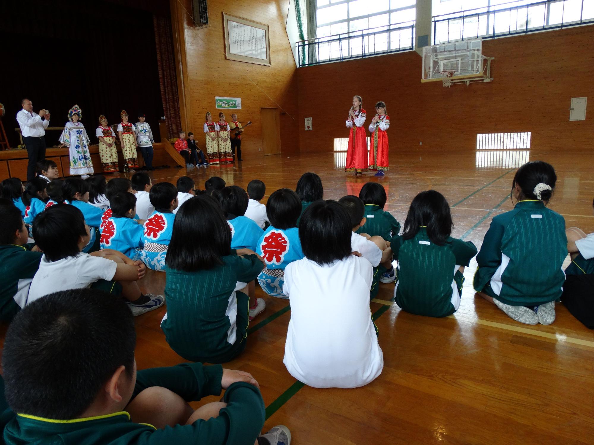 体育館で民族衣装を着た外国の子供たちが歌を披露し、それを児童たちが座って見守っている写真