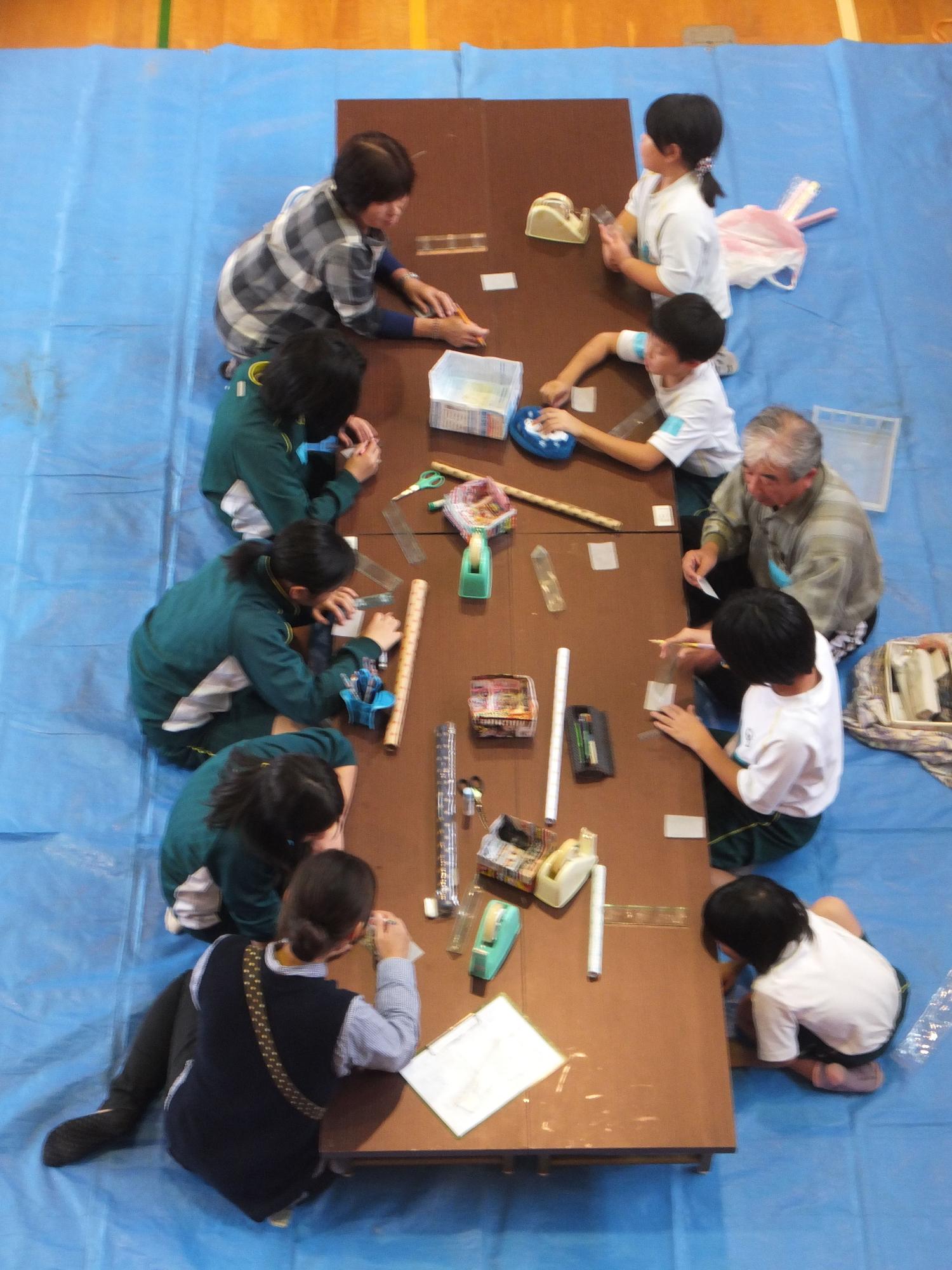 ブルーシートの上で、長机を囲んで児童たちと大人が一緒に座り、製作をしている写真