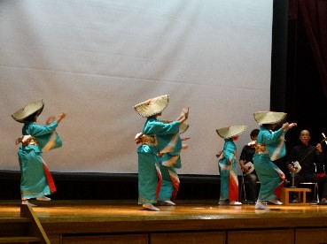 舞台で児童が笠をかぶり和装で踊っている写真