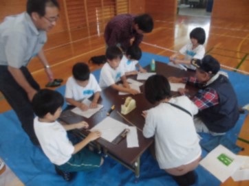体育館の机の上で児童たちが地域の講師の方と紙に書きながら作業をしている写真