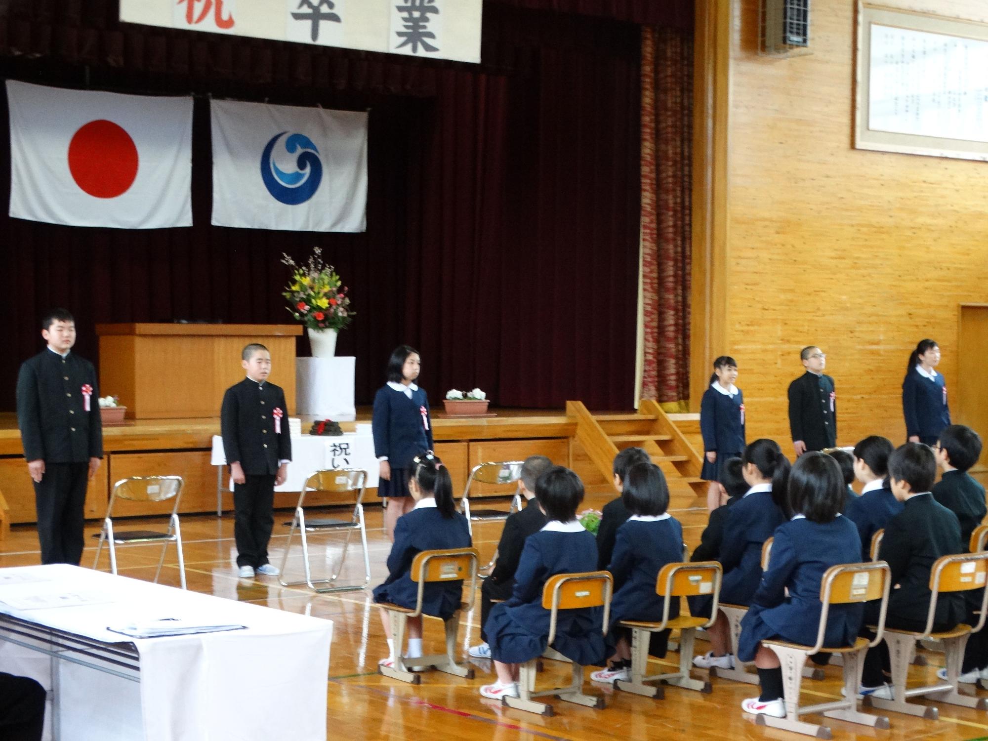 体育館で制服姿の児童が整列して立ち、それを他の児童たちが椅子に座って見ている写真