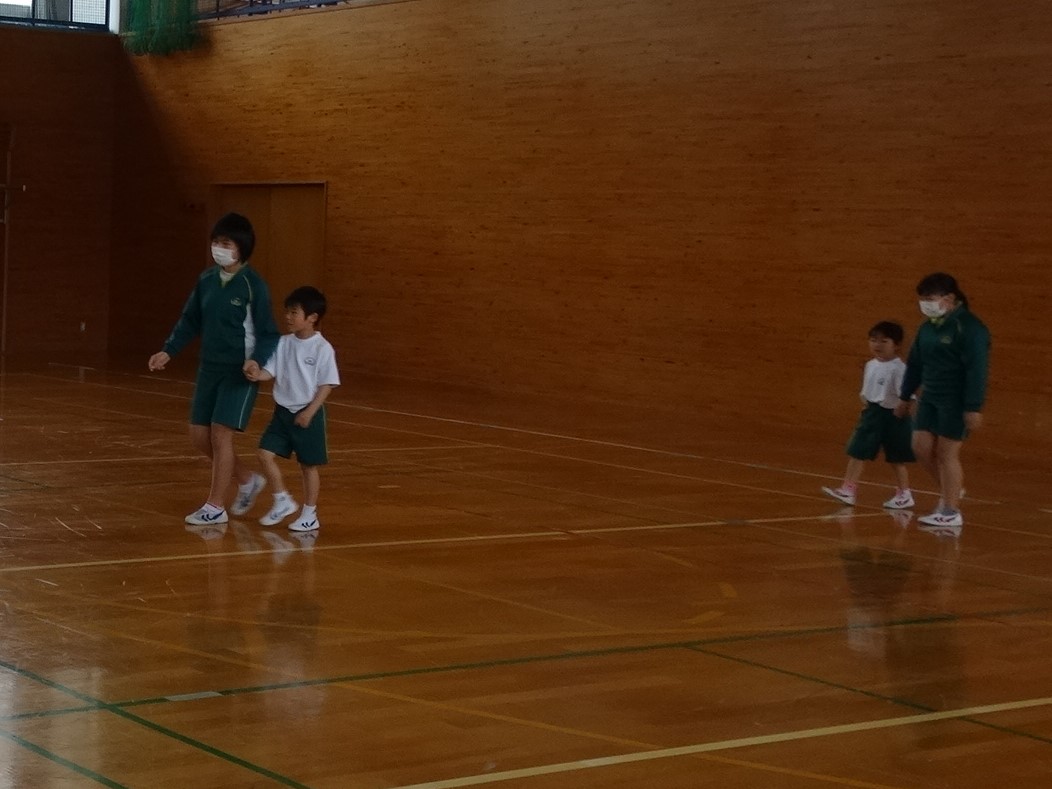 体育館の中を六年生の児童が一年生の児童と手をつなぎ一緒に歩いている写真