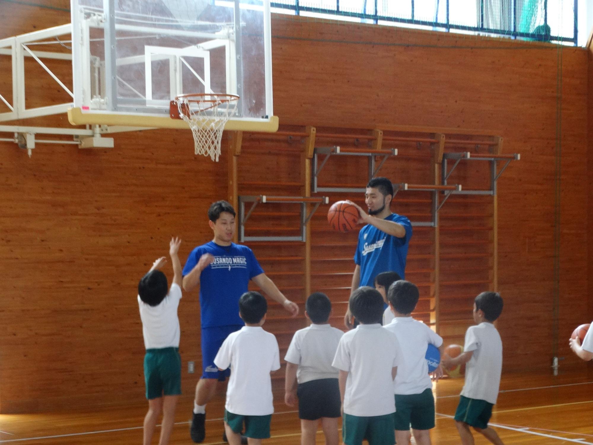 小学校の体育館でプロバスケット選手二人がシュートの時のボールの持ち方を実演しているところを真剣に聞いている児童の写真