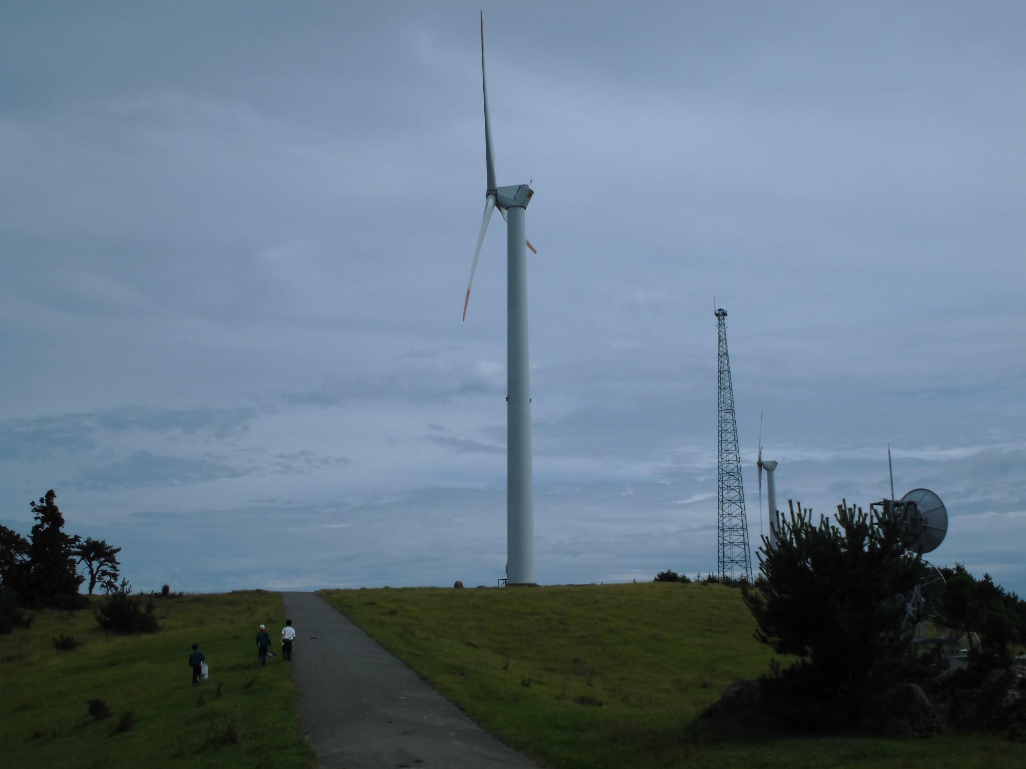 曇り空の中3人の児童が大きな風車の形をした風力発電機を観察している写真