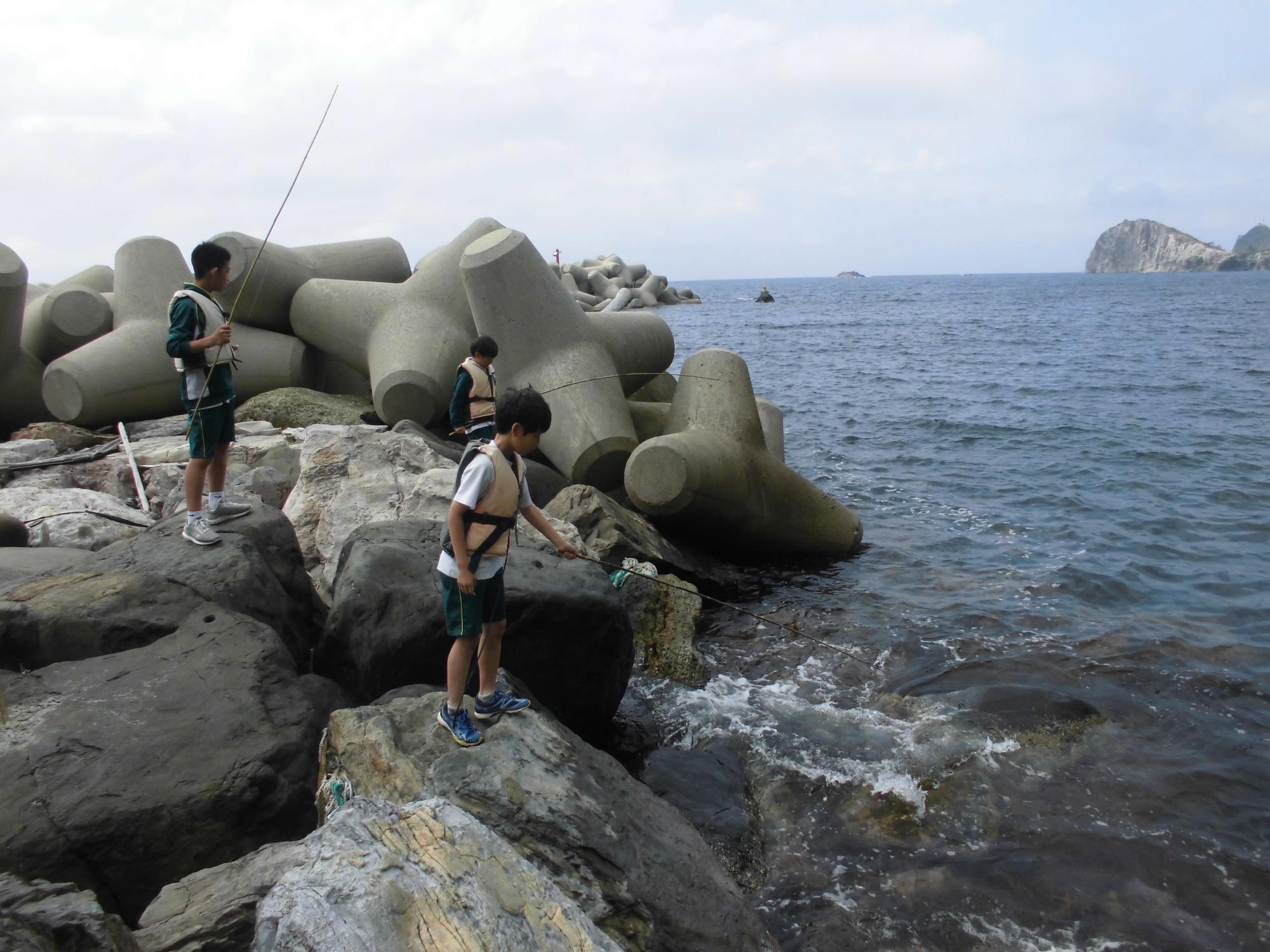 曇り空の中大きなテトラポットがある海辺で救命胴衣を着けた3人の児童が竹で作った釣り座をで釣りをしている写真