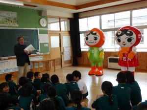 体操服姿の複数の児童が2体の着ぐるみと男性を座って見ている写真