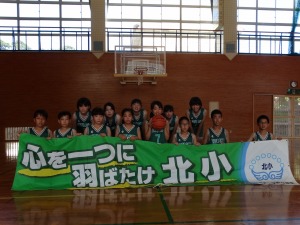 体育館で児童たちが「心を一つに羽ばたけ北小」とかかれた横断幕を掲げて整列している写真
