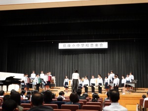 ステージ上に楽器を持った児童たちが指揮者の女性を見ながら演奏している様子を複数の観客が見ている写真