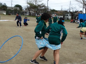 運動場で体操服姿の児童が二人一組になり、大きな布を腰に巻いて競走している写真