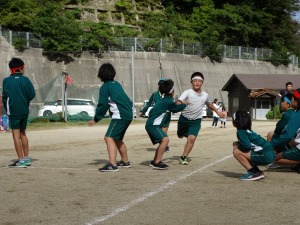 運動場で体操服姿の児童がリレーのバトンを渡している写真