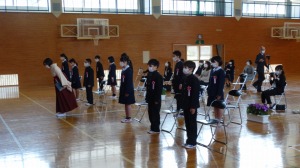 体育館で袴姿の女性がお辞儀をしている後ろに複数の児童が椅子から立ち上がり前を向いている写真