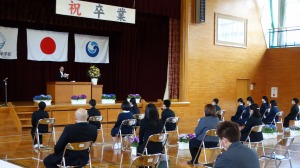 体育館のステージ上の壇上に立っている男性を複数の児童や男女が椅子に座って見ている写真