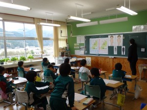 教室で男性が黒板の前に立ち、児童が机に向かって授業を受けている写真