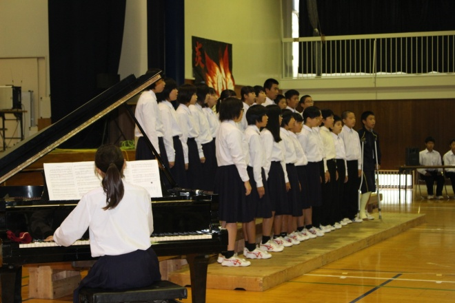 体育館で生徒が壇上で列になって立ち、その手前で1人の女子生徒がピアノを演奏している写真