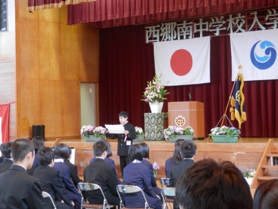 体育館のステージ手前に立っている男子生徒の話を椅子に座って複数の生徒が聞いている写真