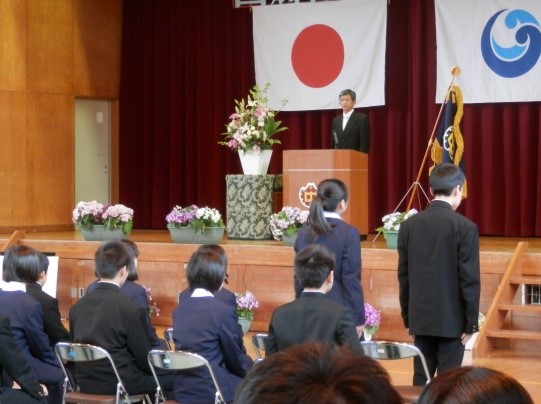 体育館のステージ上に立っている男性に向かって生徒たちが起立して立っている写真