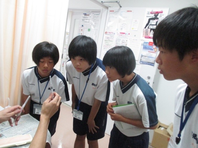 4人の生徒が注射器のようなものを見ながら説明を聞いている写真