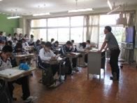 教室で生徒たちが教師の話を聞きながら机に向かっている写真