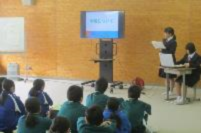 前方で生徒が資料を読み上げ、他の生徒が座って聞いている写真