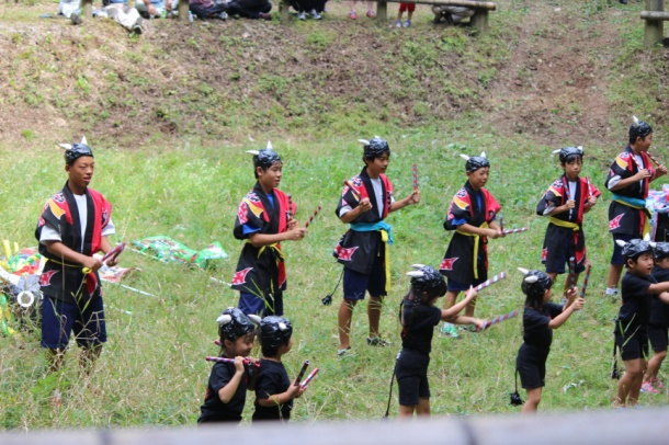 草が一面生えた場所で衣装に身を包んだ複数の生徒や園児が棒状のものを手に持ち並んでいる写真