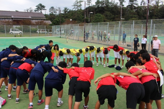 テニスコートでユニフォーム姿の生徒が肩を組みながら円陣をしている写真