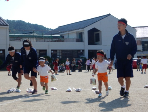 校庭でハチマキ姿の生徒が園児と手を繋いで歩いている写真