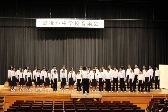 ステージ上に指揮者の方を向きながら複数の生徒たちが足を肩幅に広げて合唱している写真