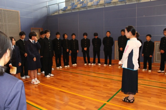 制服姿の生徒たちが円形に並び、前に立つ女性の話を聞いている写真
