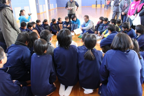 体育館で生徒たちが輪になって座り、中央の男性の話に耳を傾けている写真