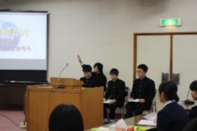 壇上で生徒が座り、一人の生徒が手を挙げて発言している写真