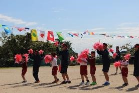 校庭で生徒と園児が一緒にポンポンを持ち踊っている写真