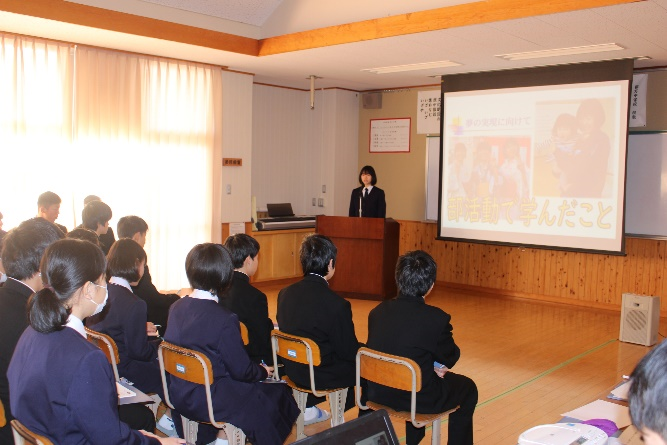 教室で一人の生徒が教壇で話をしているのを椅子に座った生徒たちが真剣に聞いている写真