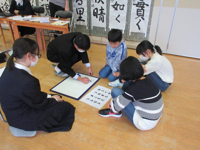 教室の床で上級生が新入生に書道を実演して見せている写真