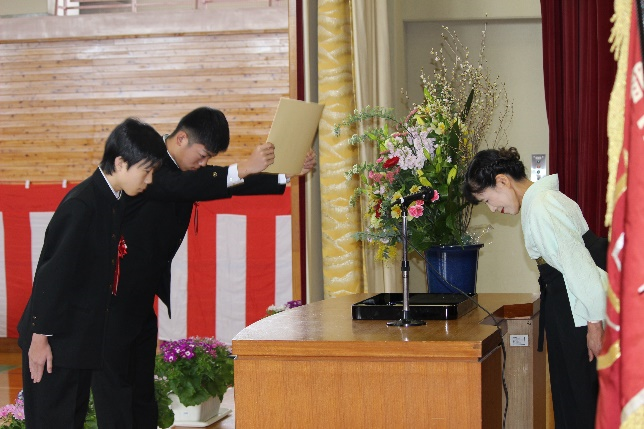 体育館で、2人の生徒が壇上で教員と向かい合い互いにお辞儀をしている写真