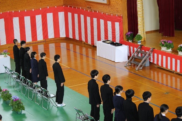 紅白の幕が張られた体育館で、卒業生全員が起立している写真