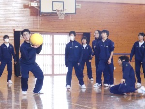 体育館で生徒が大きなボールを持ち、その様子を周囲の生徒が見守っている写真