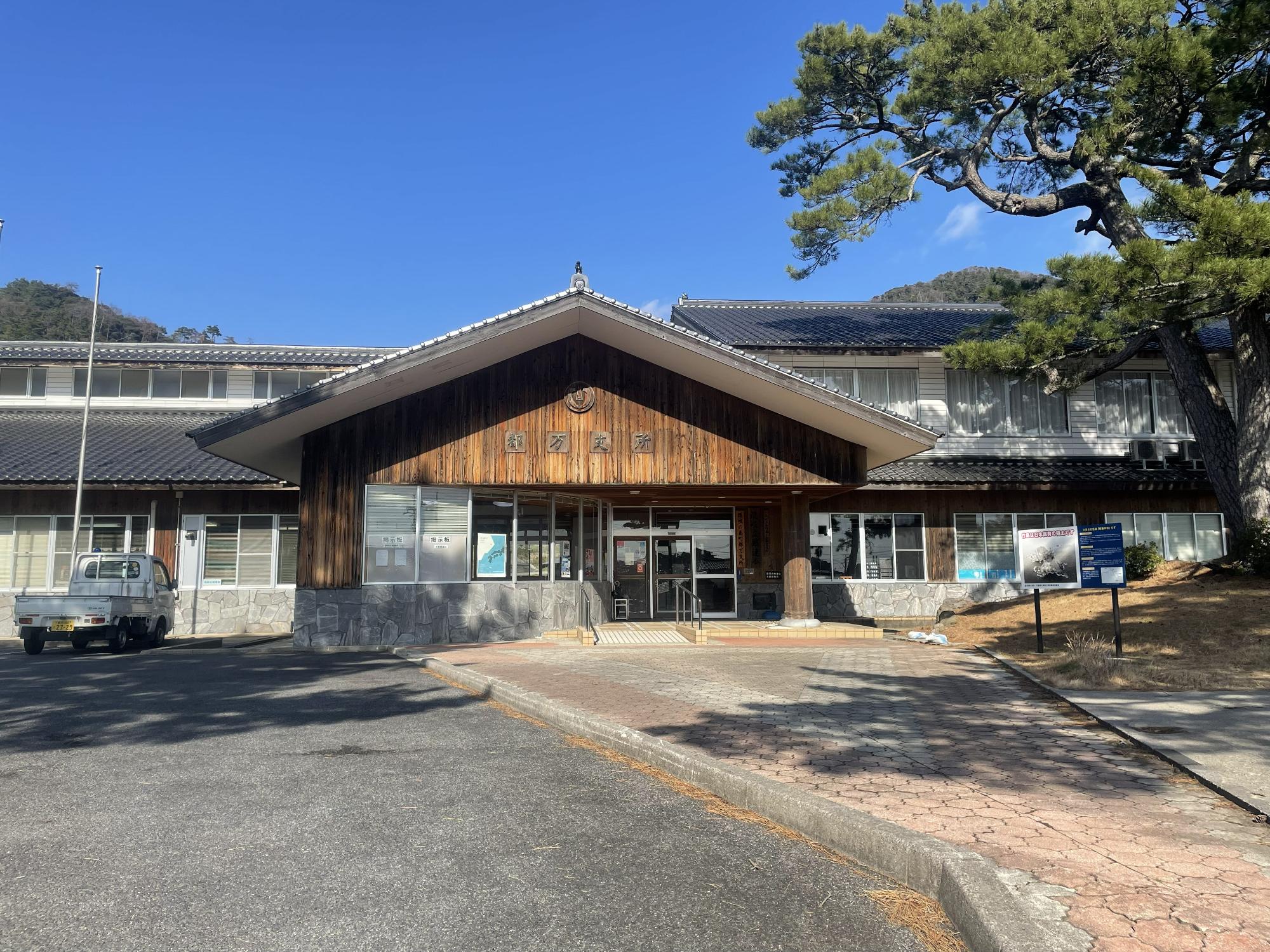 木が立っている奥に三角の屋根の平屋の建物がある写真