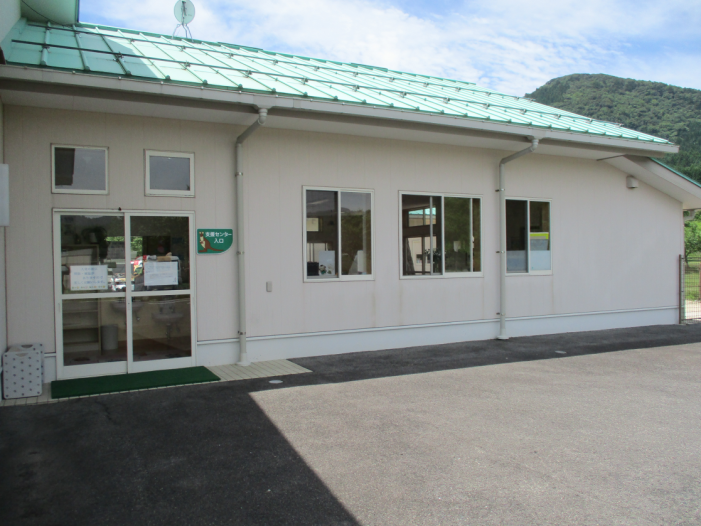 薄い青緑色の三角屋根に白い壁の建物の手前に黄土色の地面がある写真