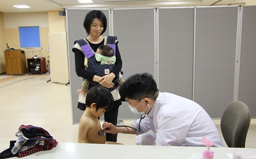 診察をしている医師と児童、それを見守る母親と赤子の写真