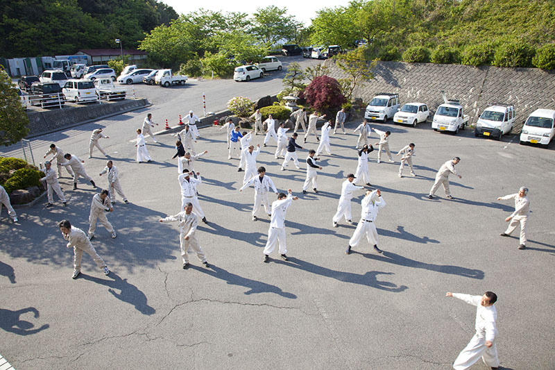 白い作業着を着てアスファルトの上で体操をしている様子の写真