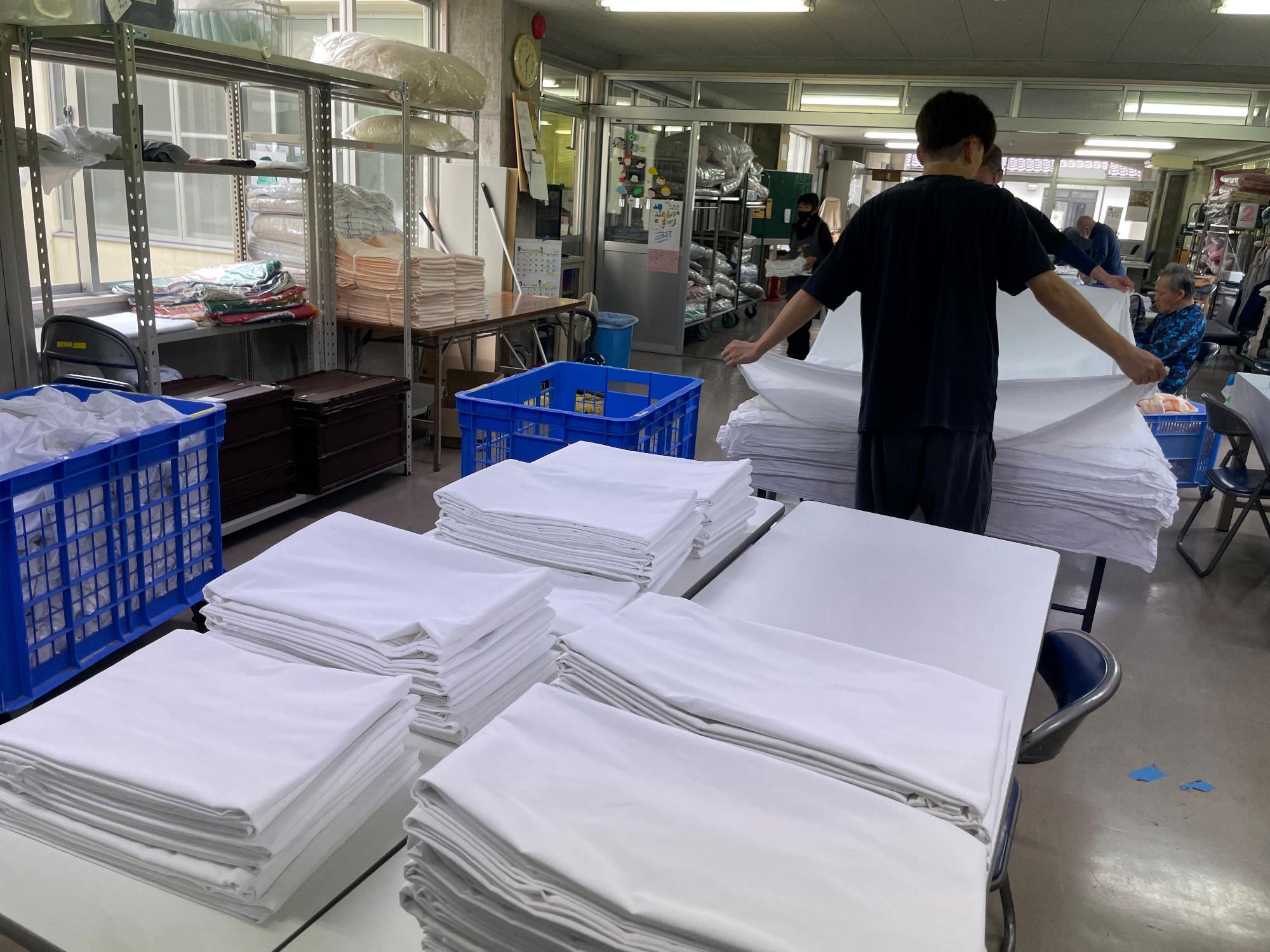 たたまれた洗濯物が机に重ねてあり、その奥で洗濯物をたたんでいる様子の写真