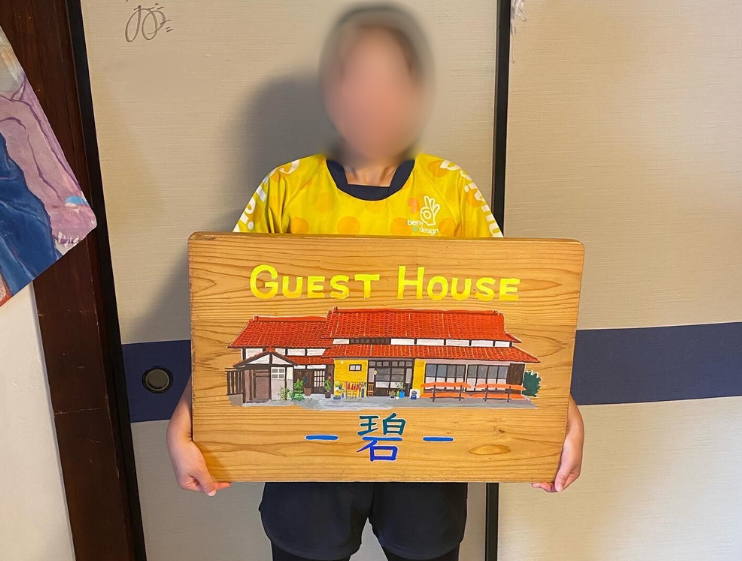 ゲストハウス碧のイラストと名前が描かれた木製の板を持って襖の前に立っている様子の写真