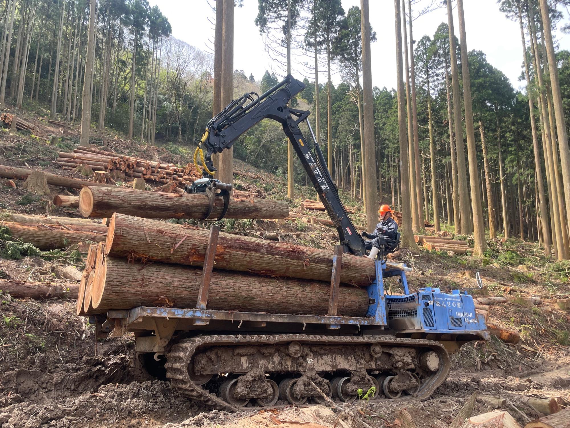クレーンのついたキャタピラの車で丸太を荷台に積んでいる様子の写真