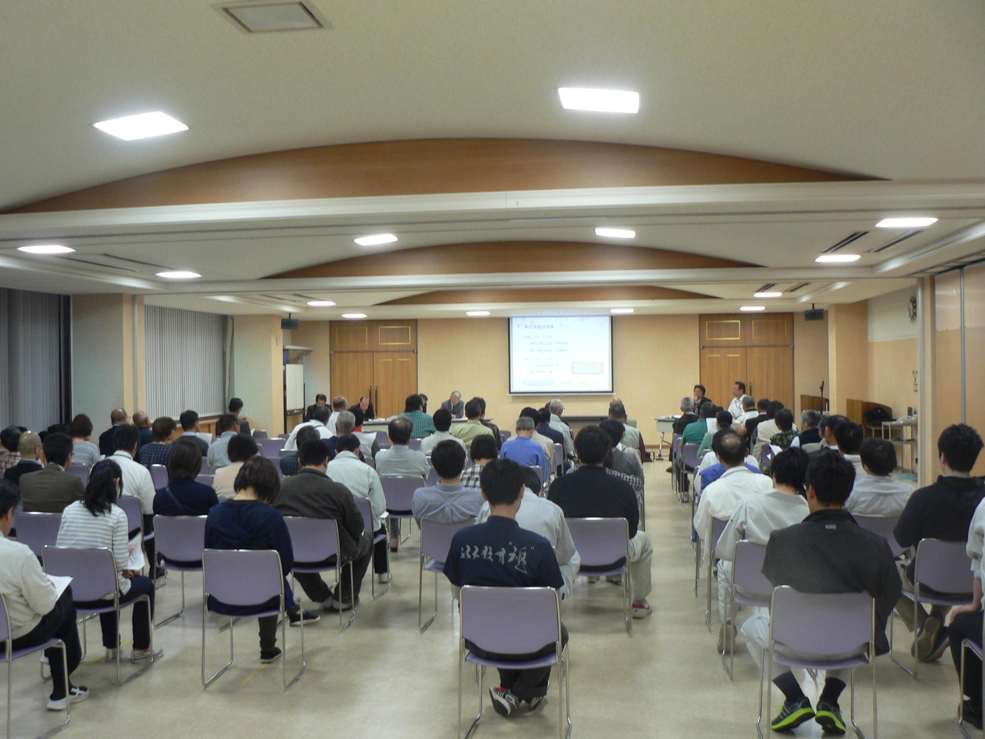 講堂に大勢の男女が集まり紫色の椅子に座り説明会に参加している写真