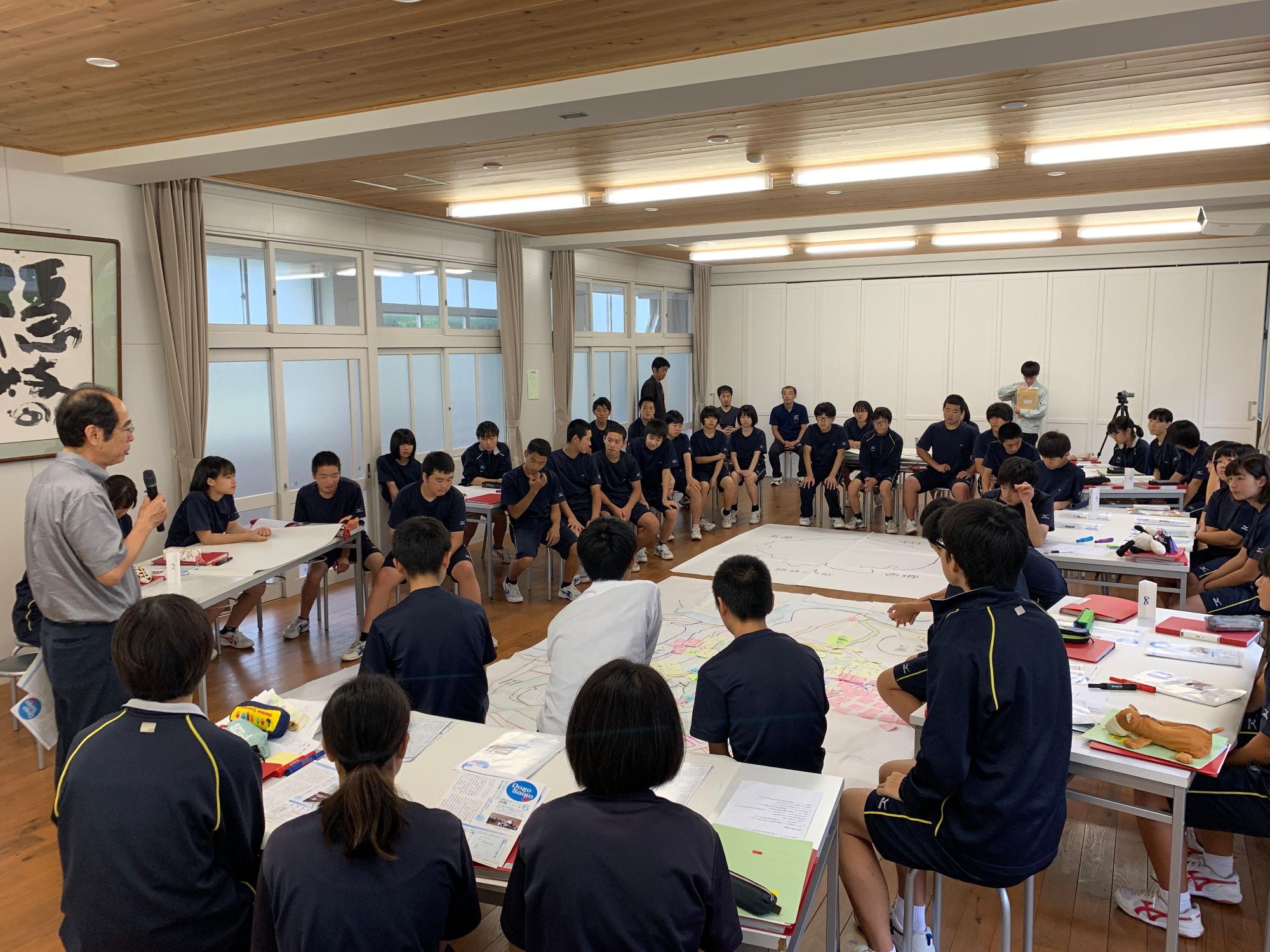 床に置かれた大きな地図を中心に座って、男性の話を聞く生徒たちの写真