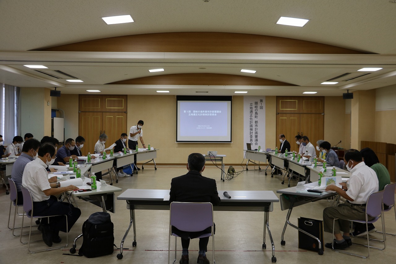 スクリーンに向かって楕円形に並んだ席に座って資料を見る人たちの写真