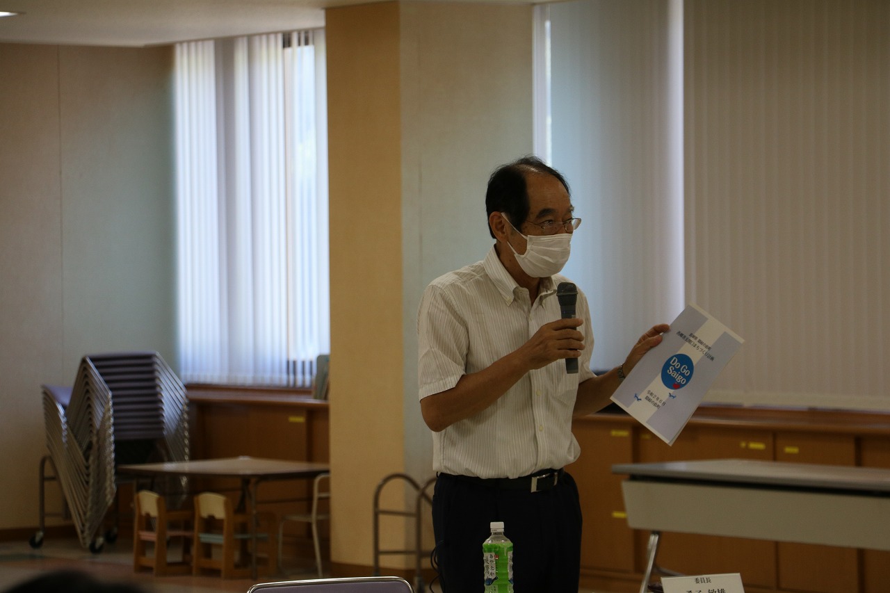 マスク姿の男性がマイクと資料を持って話している写真